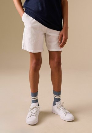 Isik kandmas valgeid põlveni ulatuvaid lühikesi pükse, tumesinist särki, hallid sokke valgete triipudega ja valgeid tenniseid, seistes ühe käega pihas.