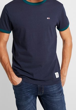 Mann trägt dunkelblaue kurzärmelige T-Shirt mit türkisfarbenem Kragen und Ärmelabschlüssen, kombiniert mit blauen Jeans, Hände in den Taschen.