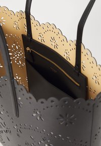 Black tote bag with perforated floral design and scalloped edges. Interior features a solid beige panel and a black zippered pocket.