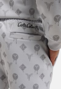 Grå joggebukser med et subtilt helmønster av logoer og symboler, med en glatt tekstur og en sidelomme for oppbevaring.