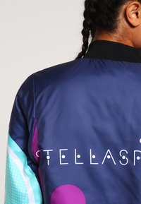 Colorful bomber jacket with navy base, patterned sleeves in turquoise and pink, featuring stylized text "STELLASPR." Smooth, glossy material.