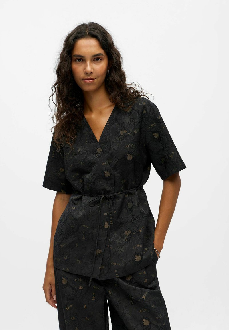 Black wrap top made of textured fabric, featuring a floral pattern in dark green and brown. Short sleeves, tie at the waist, and V-neck design.