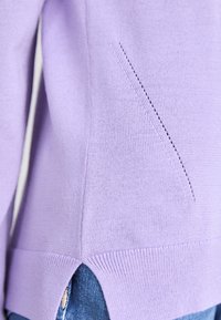 Sweater de malha lilás claro com uma racha lateral e detalhe de costura diagonal. Apresenta uma bainha canelada e textura suave. O denim é parcialmente visível.