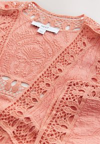 Korallinvärinen pitsipaita, jossa on monimutkaiset virkkuukuvioinnit, teksturoitu kangas ja rento malli, jossa on avonaiset yksityiskohdat reunoilla.