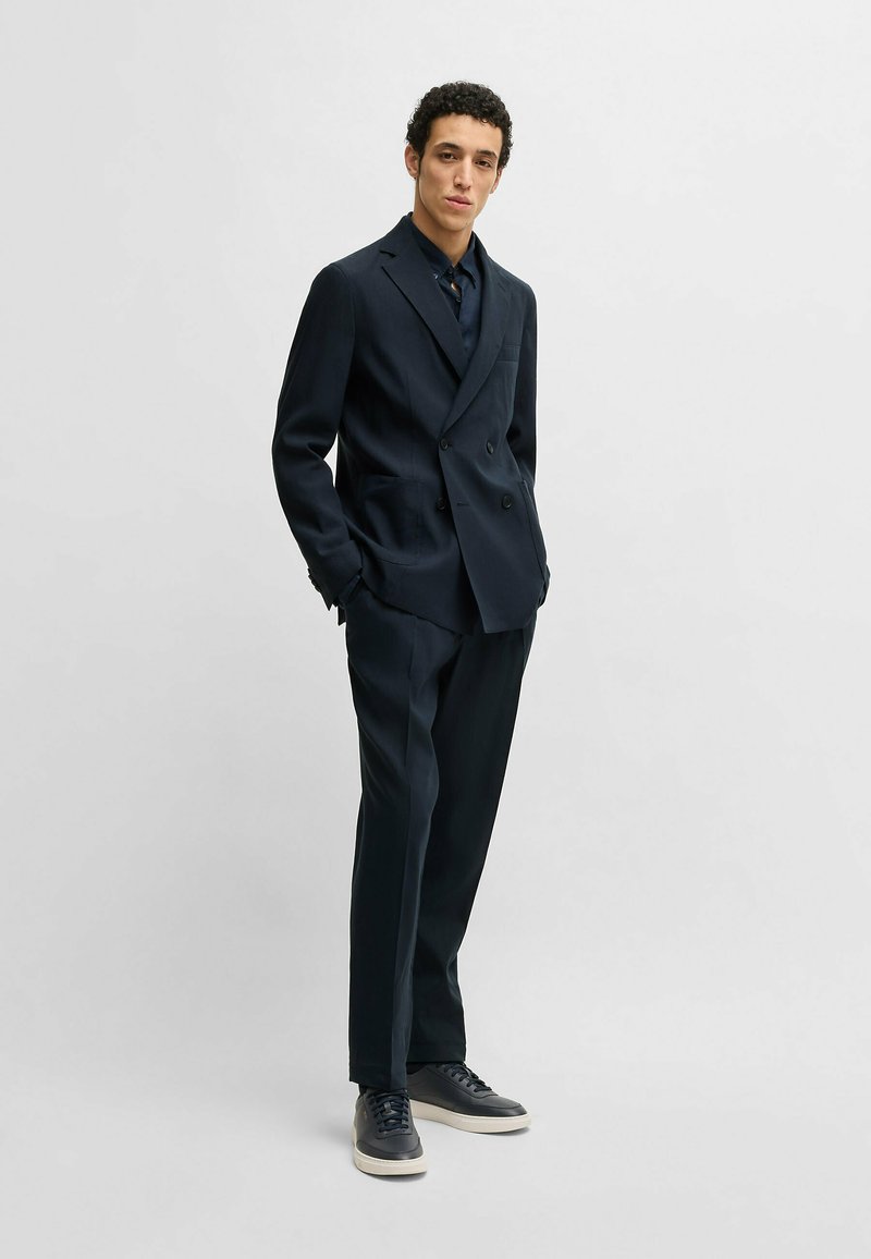 Marineblaues, schlank geschnittenes, doppelt geknöpftes Sakko mit Revers und zwei Fronttaschen, kombiniert mit dunklen Sneakers für einen modernen Look.