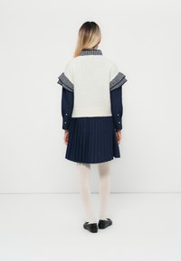 Gestrickter Elfenbeinpullover mit navy-blauen gestreiften Volantärmeln, kombiniert mit einem navy-blauen Faltenrockkleid und weißen Strumpfhosen, abgerundet mit schwarzen Ballerinas.