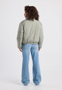 Chaqueta bomber acolchada de color verde oliva con dobladillo elástico ajustado y mangas anchas, combinada con jeans de color azul claro, ligeramente acampanados.