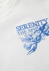 Valkoinen puuvillapusero, jossa on sininen kirjailtu teksti "SERENITY THE SPIRIT OF FASHION" yhdessä enkelimäisten hahmojen ja monimutkaisten yksityiskohtien kanssa.
