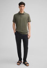 Olivgrünes Polohemd mit Kragen, drei Knöpfen und kurzen Ärmeln, kombiniert mit schwarzen Hosen und schwarzen Sandalen mit Schnallen-Design.