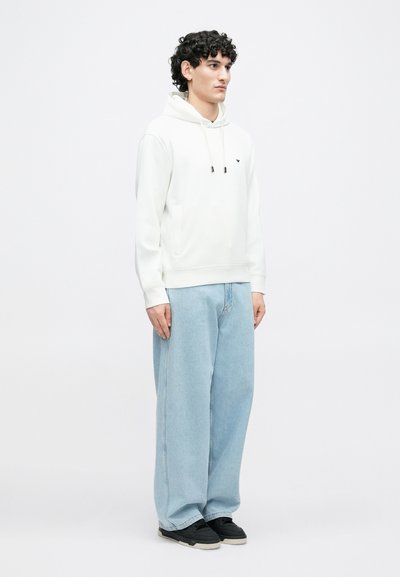 Balta džemperio su kapišonu, reguliuojamomis virvutėmis ir priekyje esančia kišene, derinamas su šviesiai mėlynais plačiomis kelnėmis ir juodais sportbačiais, minimalistinis prekinis ženklas.