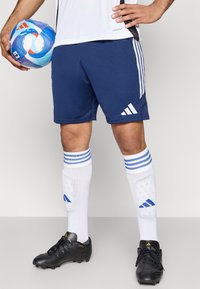 Ναυτικά σορτς ποδοσφαίρου με το λευκό λογότυπο της Adidas, συνδυασμένα με λευκές κάλτσες που έχουν μπλέ λωρίδες και μαύρα παπούτσια ποδοσφαίρου, κρατώντας μια πολύχρωμη μπάλα ποδοσφαίρου.