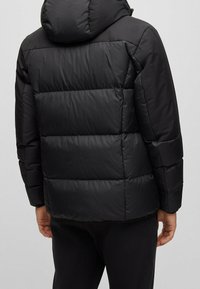 Svart puffjacka med ett kviltat mönster, som har huva, elastiska ärmslut och en slät yta. Visad från bakvyn.