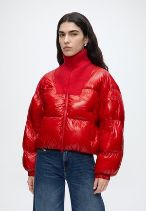 Vrouw met lang donker haar die een glanzend rode pufferjas met hoge kraag en blauwe spijkerbroek draagt, staand tegen een effen witte achtergrond.