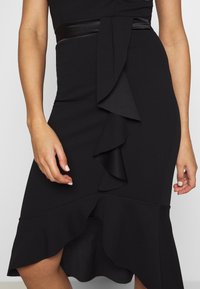 Svart klänning med en volangkant i asymmetrisk stil och en välsittande silhuett, som har ett glansigt svart midjeband för kontrast. Texturerat tyg.