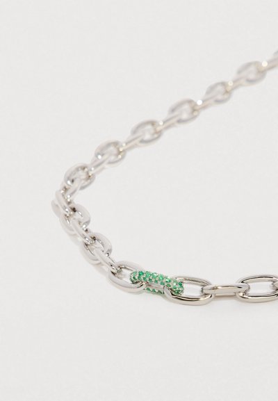 Srebrny naszyjnik z łańcuszkiem, którego pojedynczy ogniwo jest ozdobione małymi zielonymi kamieniami szlachetnymi na białym tle.