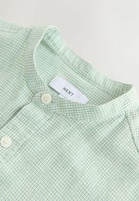 Chemise vert clair à motif quadrillé, avec un col à revers, deux boutons, et une étiquette en tissu portant la marque "NEXT".