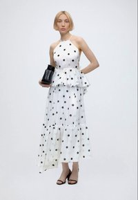 Femme en robe blanche sans manches à pois noirs, tenant un sac noir texturé et portant des talons ouverts noirs.