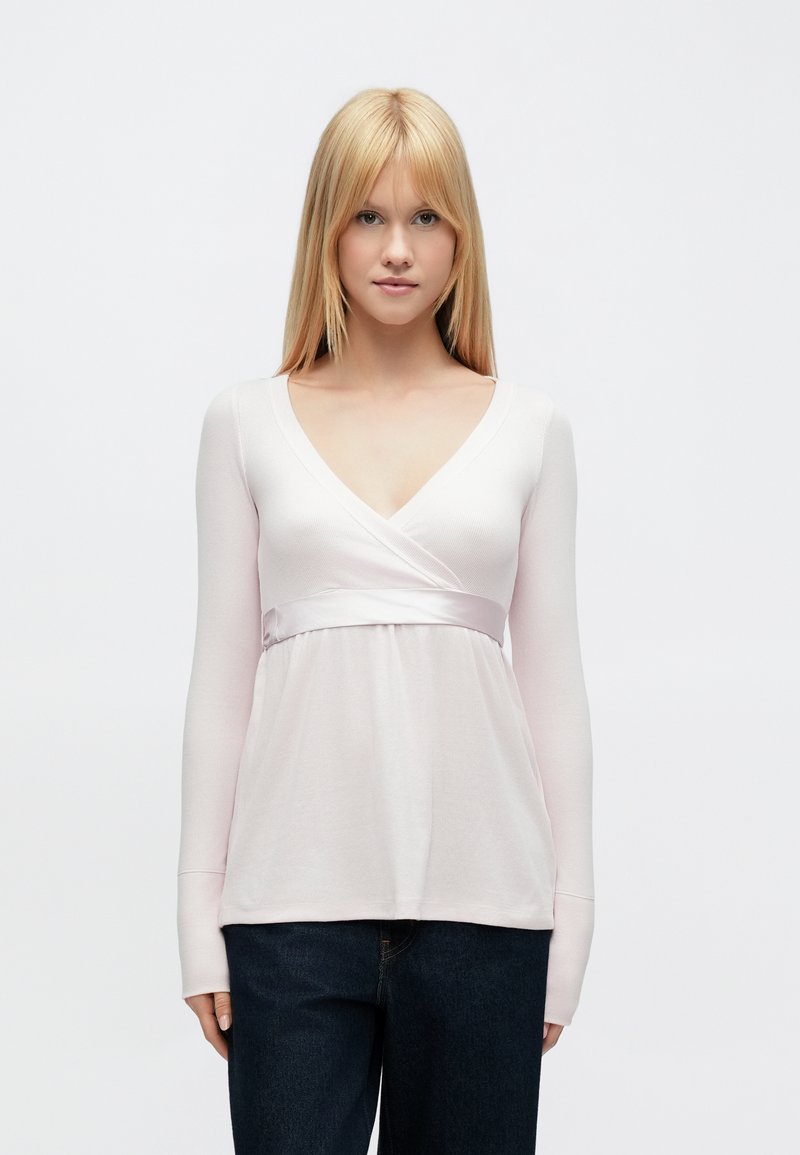 Világos rózsaszín hosszú ujjú felső V-nyakkivágással, átkötős kialakítással és szatén derékpánttal. Puha, texturált anyagból készült. Sötét farmerrel párosítva.