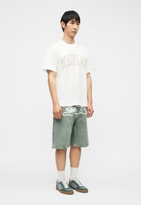 Νέος άνδρας που φοράει λευκό γραφικό μπλουζάκι, τζιν σορτς με λευκό σχέδιο καρτούν, λευκές κάλτσες και τιρκουάζ αθλητικά παπούτσια, κοιτάζοντας ελαφρώς προς τα δεξιά.