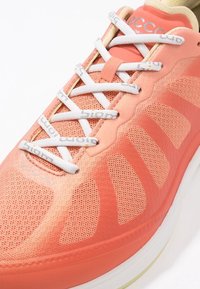 Chaussure de sport rose corail avec des lacets blancs, une tige en mesh respirant et des coutures détaillées sur les côtés et la zone des orteils.