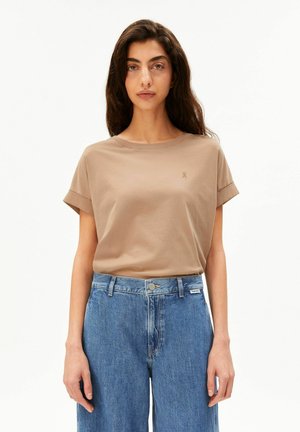 Vrouw met lang donker haar die een beige T-shirt met korte mouwen en een high-waisted blauwe spijkerbroek draagt, staand tegen een effen witte achtergrond.