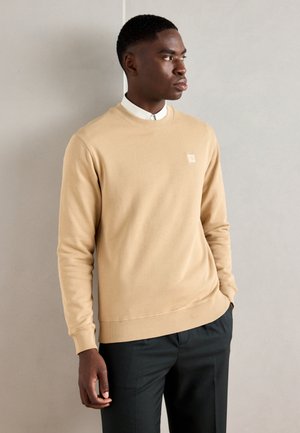 Beige sweatshirt with ribbed cuffs and hem, round neck design, featuring a small logo patch on the chest. Paired with dark trousers.