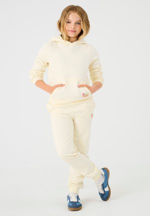 Gele hoodie met zak aan de voorkant en bijpassende broek, voorzien van subtiele branding. Model draagt blauwe sportschoenen met witte accenten.