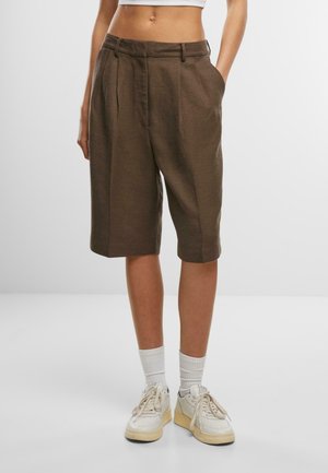 Brown pleated shorts made of textured fabric. Features side pockets, a waist belt, and a relaxed fit, paired with white socks and sneakers.