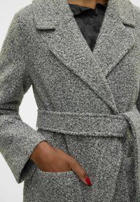 Manteau gris texturé avec une taille ceinturée, des revers larges et deux poches avant ; il est réalisé en un matériau doux en tricot bouclé.