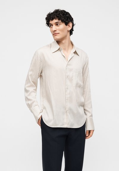Νέος άντρας με κατσαρά μαλλιά φοράει μια κρεμ ριγωτή μπλούζα με μακριά μανίκια και σκοτεινό παντελόνι, στέκεται με το ένα χέρι στην τσέπη και κοιτάζει αριστερά.