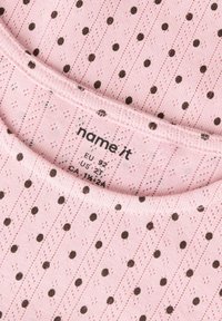 Jasnoróżowa tkanina o fakturowanym wzorze, z brązowymi groszkami i ukośnymi paskami. Okrągły dekolt z metką wskazującą rozmiar i markę.