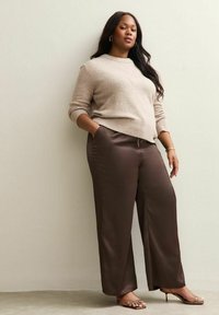 Beige knit sweater with ribbed hem paired with brown satin wide-leg pants. The model stands with hands in pockets, wearing strappy nude heels.