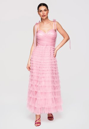 Femme en robe en tulle rose sans manches à volants avec bretelles nouées aux épaules et sandales à talons hauts magenta à lanières, posant devant un fond blanc.