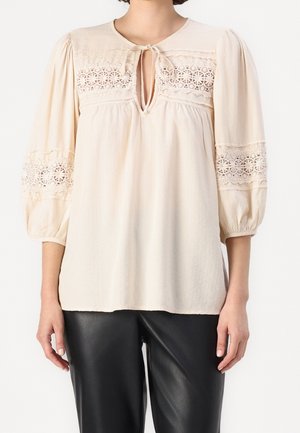 Blouse beige avec détails en dentelle sur la poitrine et les manches, fermeture par lien au niveau de l'encolure en forme de goutte, associée à un pantalon en cuir noir.