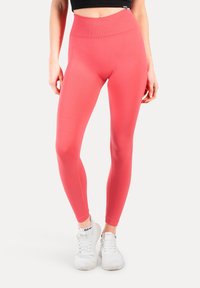 Persoon die fel koraalkleurige leggings met hoge taille en witte sportschoenen draagt, tegen een effen witte achtergrond.