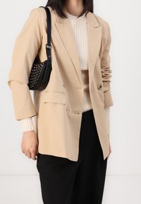 Femme portant un blazer beige sur un pull crème côtelé et un pantalon noir, avec un sac à bandoulière noir clouté sur son côté gauche.