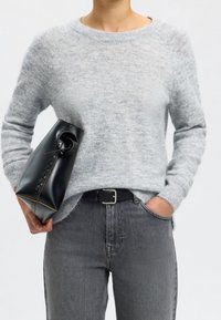 Personne portant un pull gris clair et un jean gris foncé, tenant une pochette en cuir noir dans une main, l'autre main dans la poche.