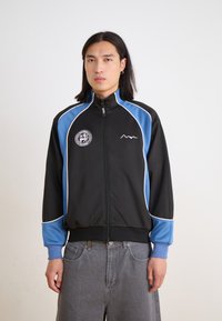 Black athletic jacket with blue accents and white piping. Features a high collar, full zipper, and embroidered logo on the chest.