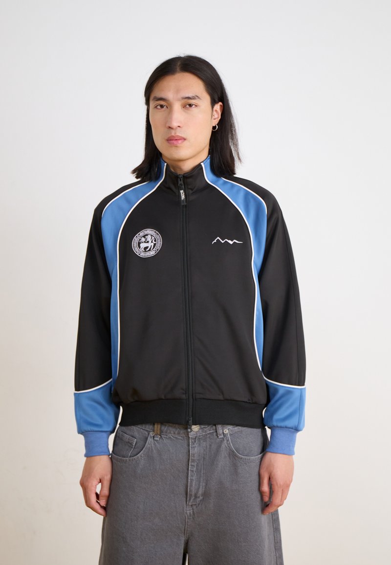 Black athletic jacket with blue accents and white piping. Features a high collar, full zipper, and embroidered logo on the chest.
