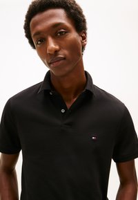 Giovane uomo con capelli corti e ricci che indossa una polo nera con un piccolo logo rosso, bianco e blu sul petto sinistro, su uno sfondo semplice.