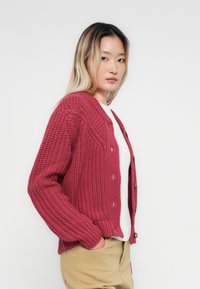 Νεαρή γυναίκα με ξανθά ομπρέ μαλλιά, φορώντας μια κόκκινη πλεκτή ζακέτα πάνω από ένα άσπρο τοπ και μπεζ παντελόνι, στέκεται στο πλάι με τα χέρια στις τσέπες.