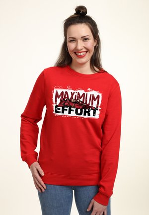 Žena se usmívá a má na sobě červenou mikinu s nápisem „Maximum Effort“ a grafikou superhrdiny v ležící poloze, doplněnou modrými džíny.
