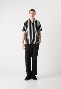 Chemise à rayures noires et blanches à manches courtes avec un col, une poche, portée avec un pantalon noir ample et des chaussures noires.