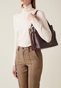 Braune Lederhandtasche mit goldenen Beschlägen, die ein strukturiertes Design und zwei Seitentaschen aufweist, gehalten von einer Person, die einen cremefarbenen Rollkragenpullover trägt.