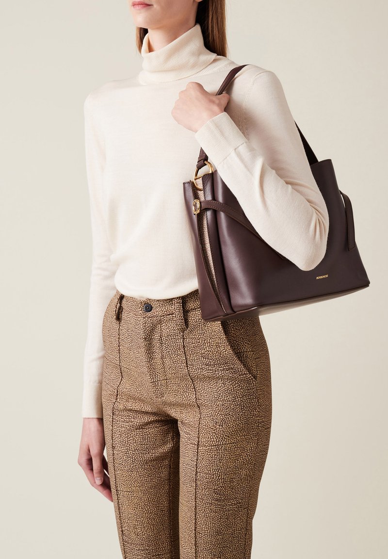 Braune Lederhandtasche mit goldenen Beschlägen, die ein strukturiertes Design und zwei Seitentaschen aufweist, gehalten von einer Person, die einen cremefarbenen Rollkragenpullover trägt.