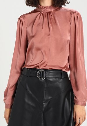 Kvinde iført en satinbluse med lange ærmer og høj hals i støvet pink, stoppet ned i en sort lædernederdel med et sølvfarvet bælte med dobbelt ring.