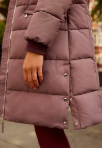 Puffermantel i dämpad mauve med kviltad textur, silverdragkedjor och tryckknappar. Ärmen har en mörkröd ribbad mudd.
