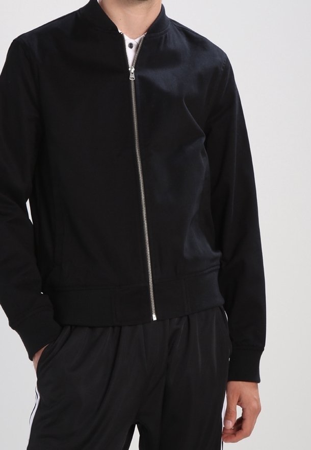 Homme portant une veste noire zippée et un pantalon noir avec des bandes blanches sur les côtés, debout avec une main dans la poche devant un fond uni.