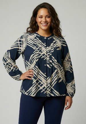Femme aux cheveux bouclés foncés portant un chemisier bleu marine avec des lignes abstraites beiges et un pantalon bleu marine, souriante, une main sur la hanche.