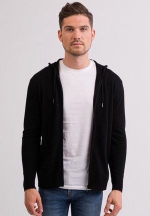 Sort zip-up hættetrøje med en blød tekstur, slim fit og to forlommer. Båret over en hvid t-shirt og parret med blå jeans.